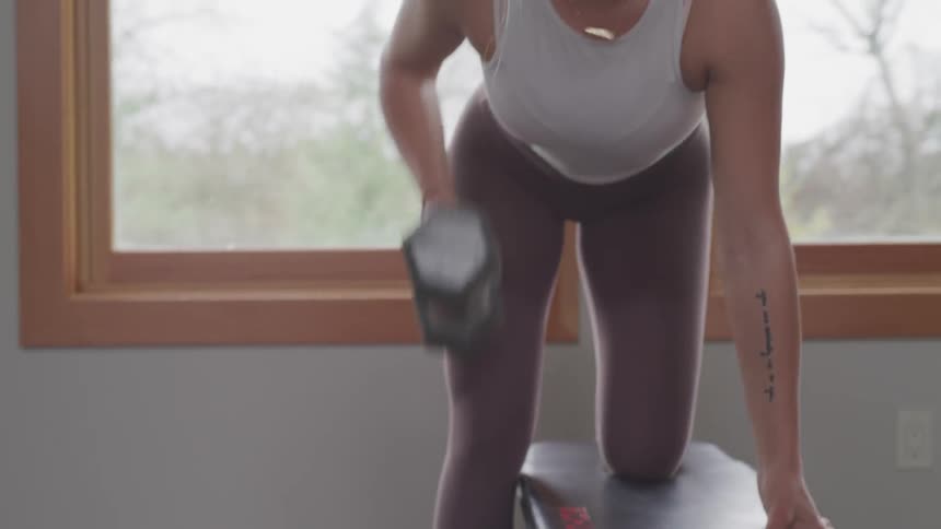 A woman performs one arm dumbbell rows on a flat bench near a window