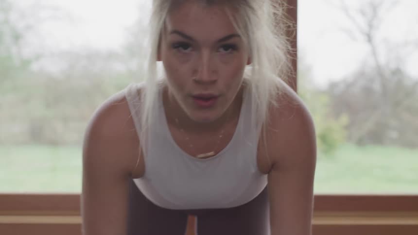 A blonde female is bent over doing triceps exercises with a free weight in front of a window
