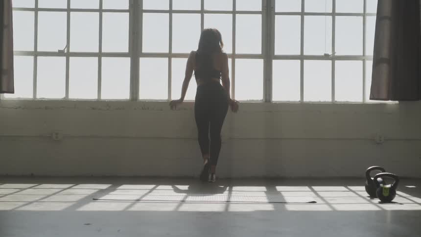 A woman in fitness apparel stands in a well lit studio at a window as there is a yoga mat and kettle bell weights on the floor nearby