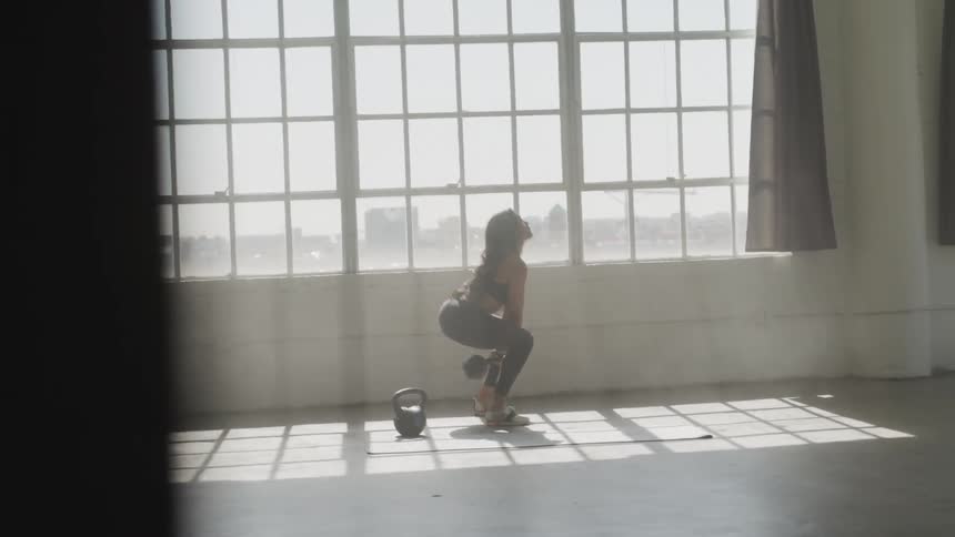 A woman in athletic apparel is doing overhead kettle bell swings near a sunny window