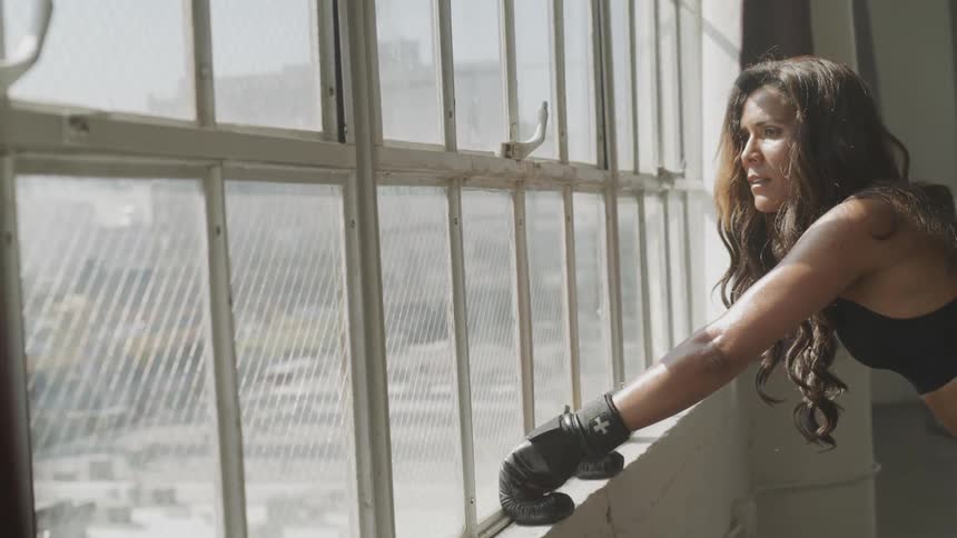 A woman wearing boxing gloves pauses to look out a window
