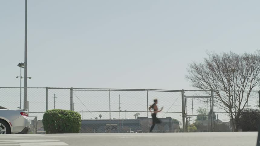 A woman wearing workout gear runs past a fence and parked car near an athletic field