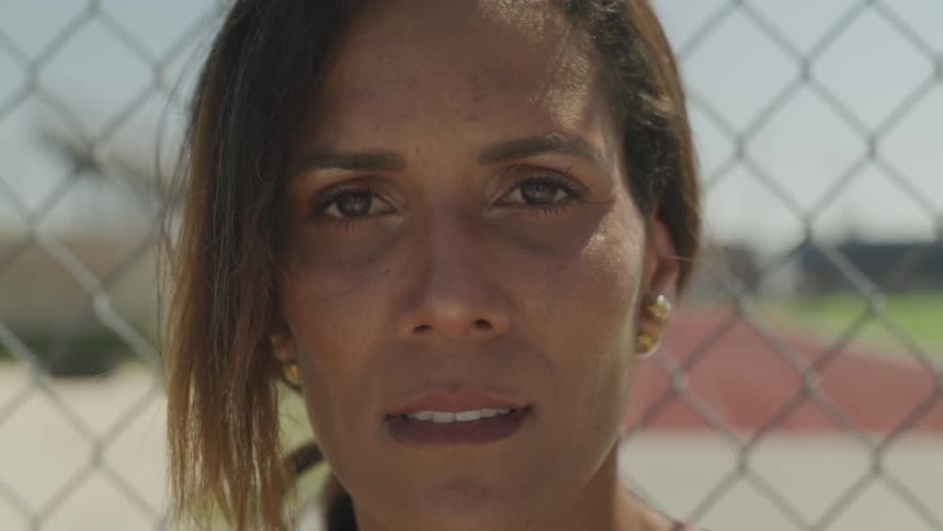 A woman stands in front of a chain link fence and looks forward with an attentive gaze