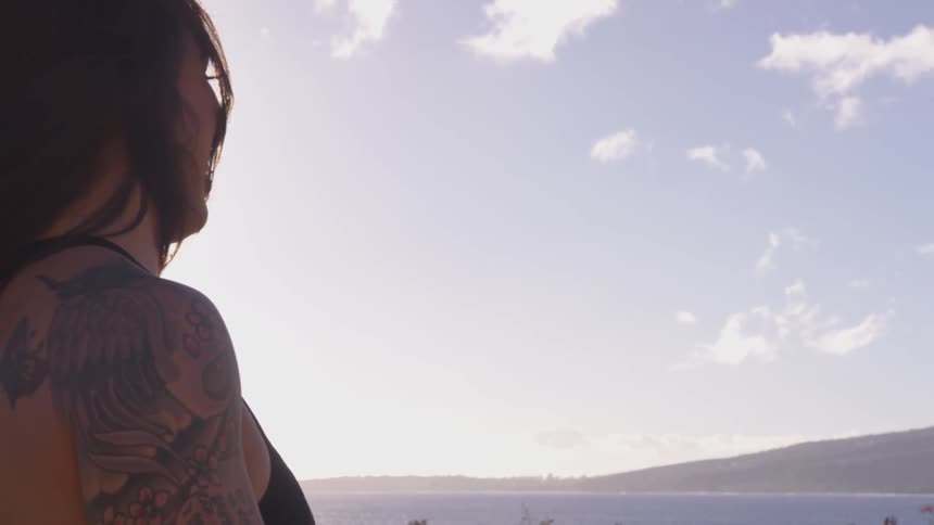 Brown haired woman with blowing hair and tattoo on her arm lifts weight outside in the sun
