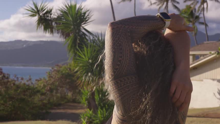 A woman has her hands stretched above her head to behind her back looking towards a body of water