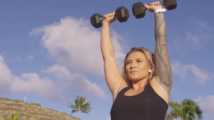 A tattooed woman lifting dumbbells outdoors on a sunny day