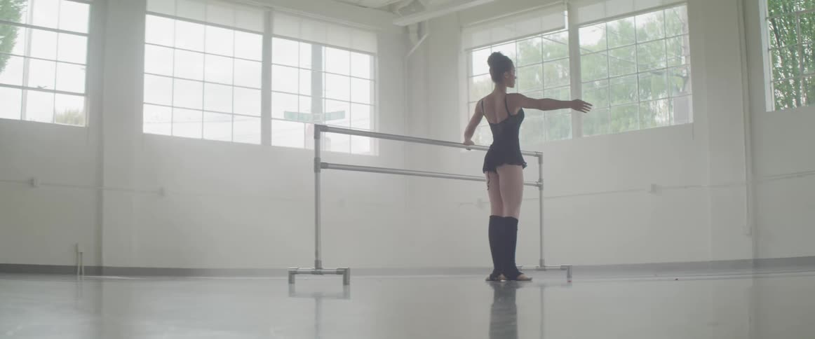 A ballerina performs a pli and touches her toes in a dance studio with a shiny floor and white walls