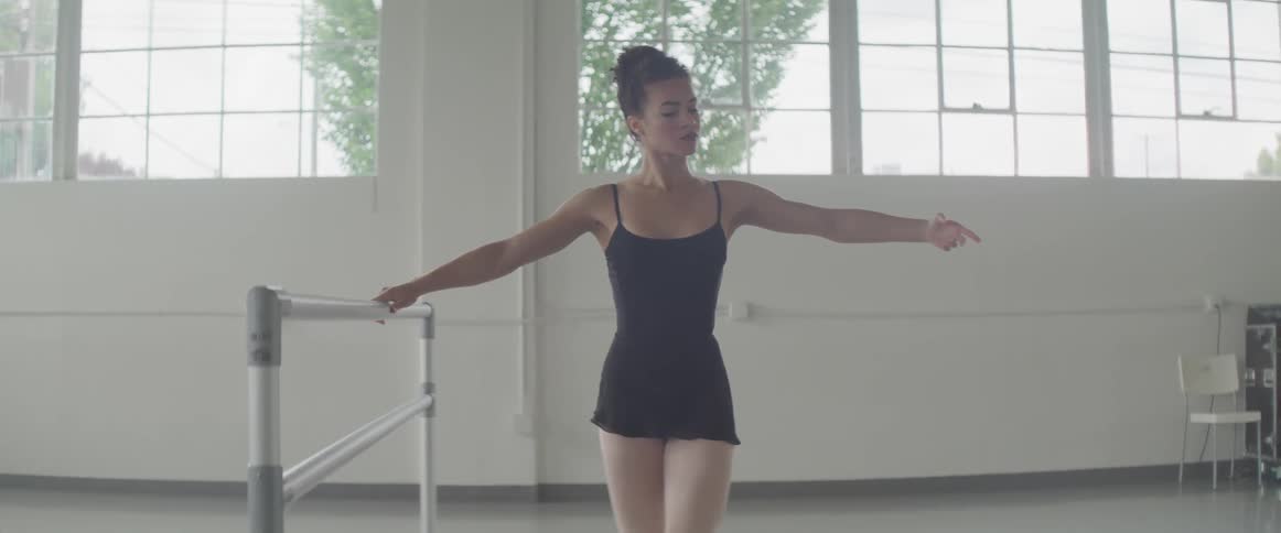 A female ballerina is performing basic dance warm up moves holding onto a barre in a dance studio