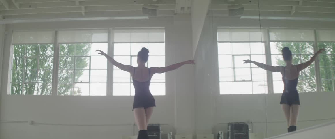 A female ballerina is dancing gracefully in front of a mirror in a dance studio with large windows
