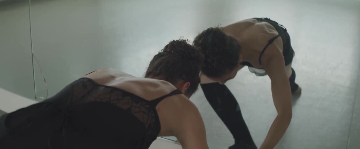 A dancer in a black leotard practices moves in front of a mirror in a large dance space full of windows