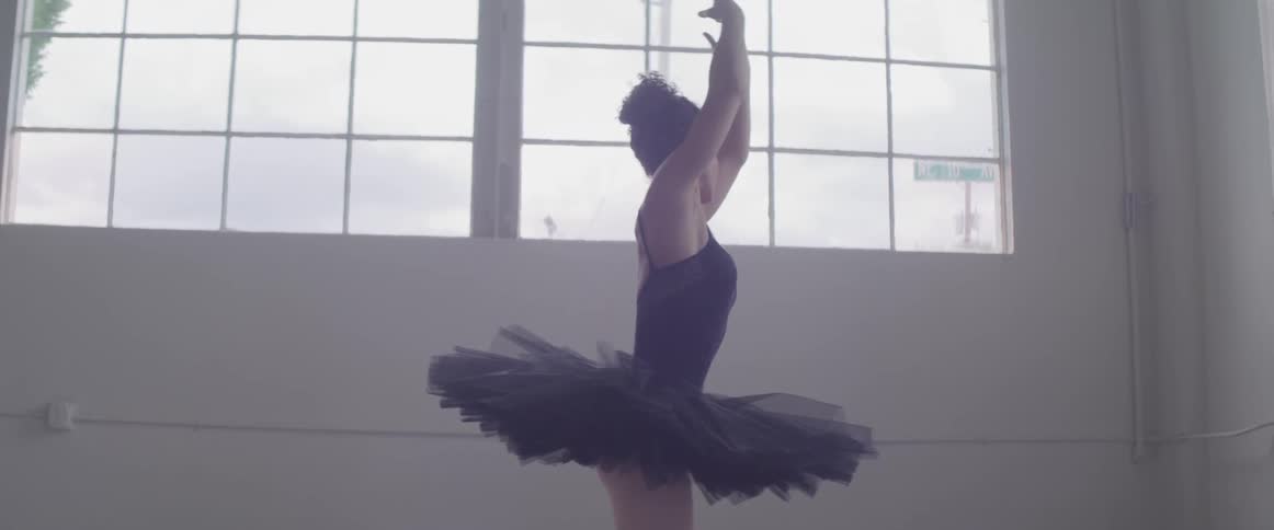 A ballerina in a black tutu dancing in a white room before two large windows during the day