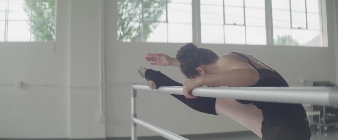 Dancer in black leotard exercises on bar in front of windows