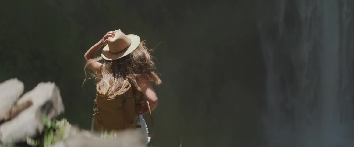 A woman in denim shorts vest and a hat stands in front of a waterfall