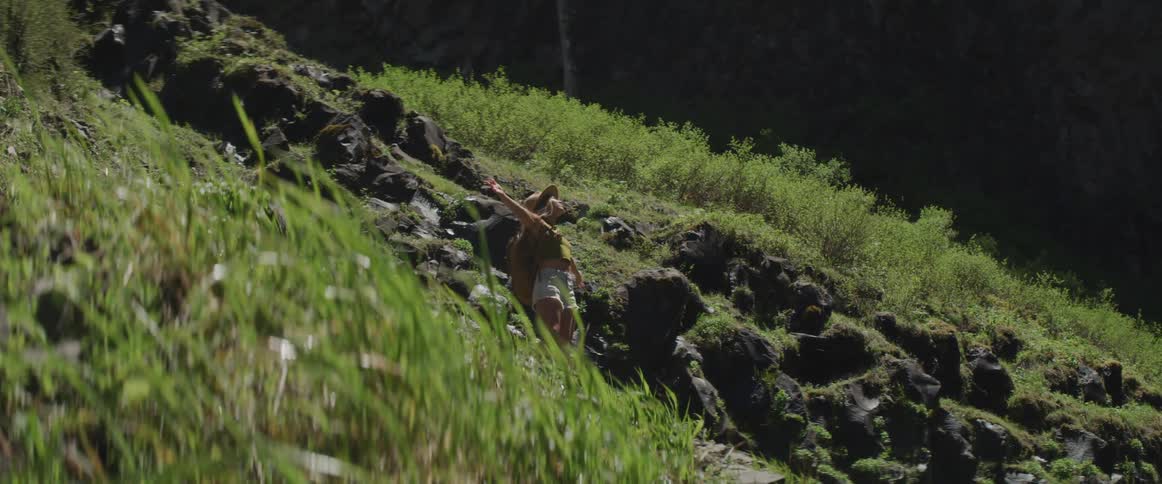 A woman in denim shorts stands on a hillside path and stretches her arms out into the wind