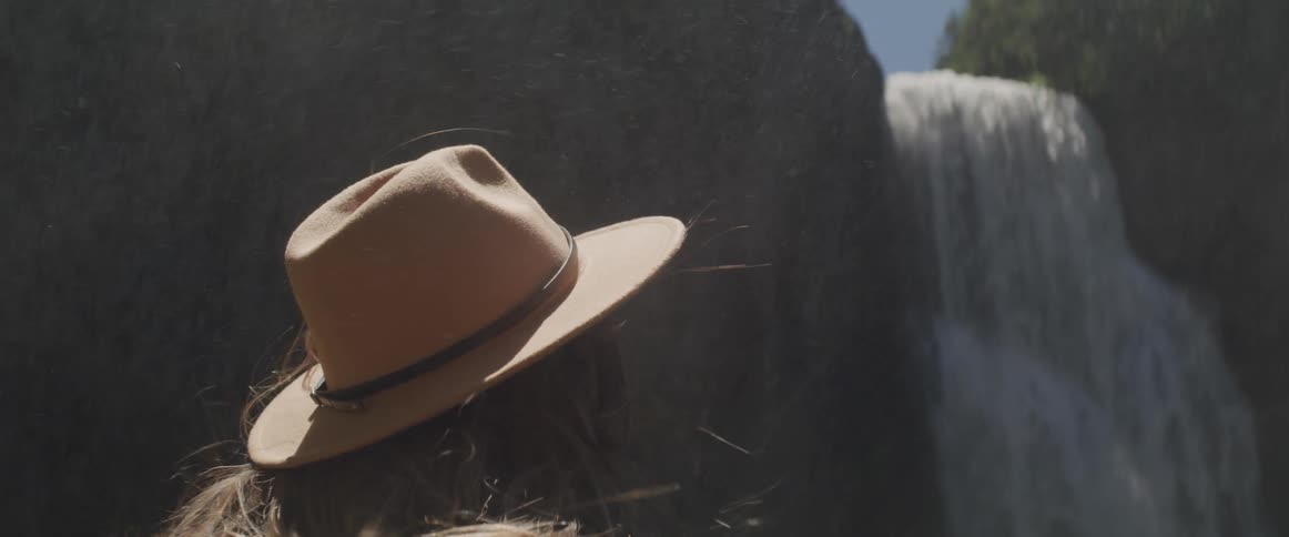 Woman wearing wide hat looks at the waterfall on a sunny day