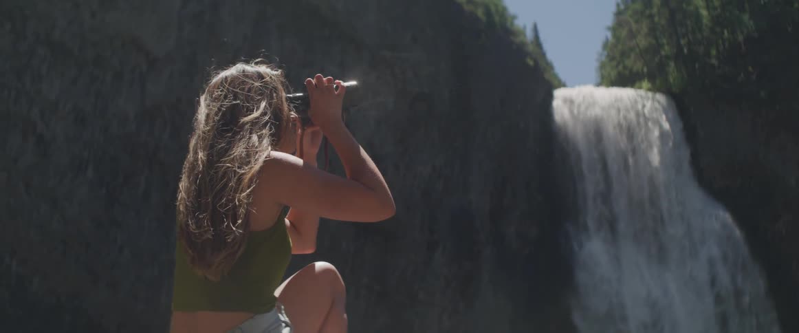 Woman l looks around through binoculars next to the waterfall on a sunny day
