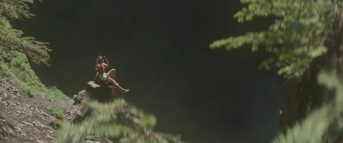 A female in denim shorts sits on a tree stump and looks out of binoculars