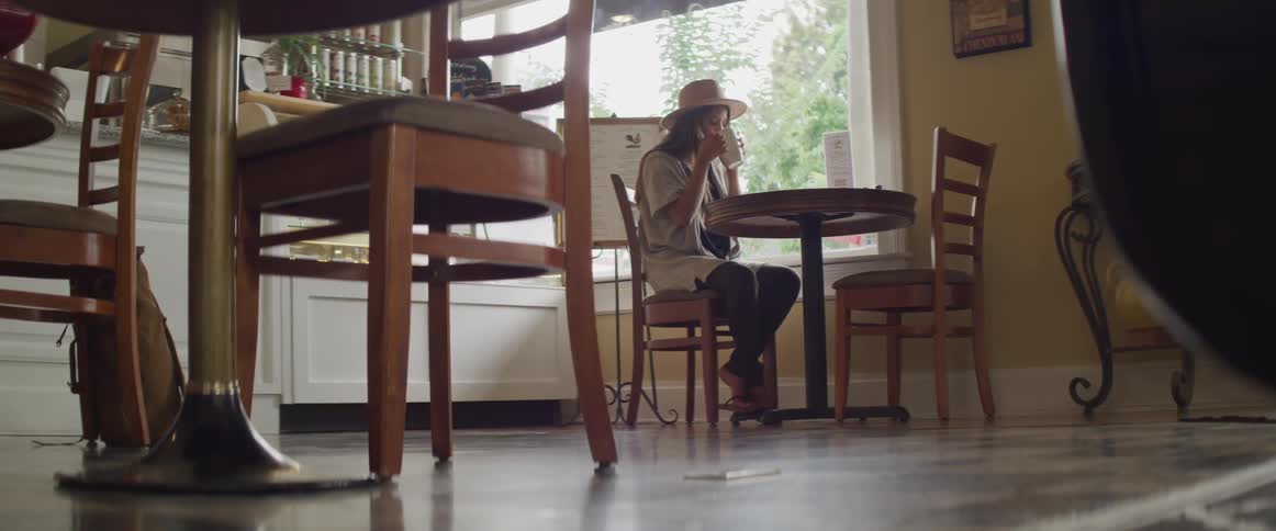Woman wearing a straw hat is sitting at a round table in a small town bakery while sipping coffee from a white mug and looking out the window