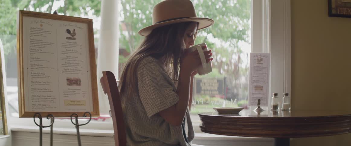 A woman sitting at a table in a cafe drinking from a large mug