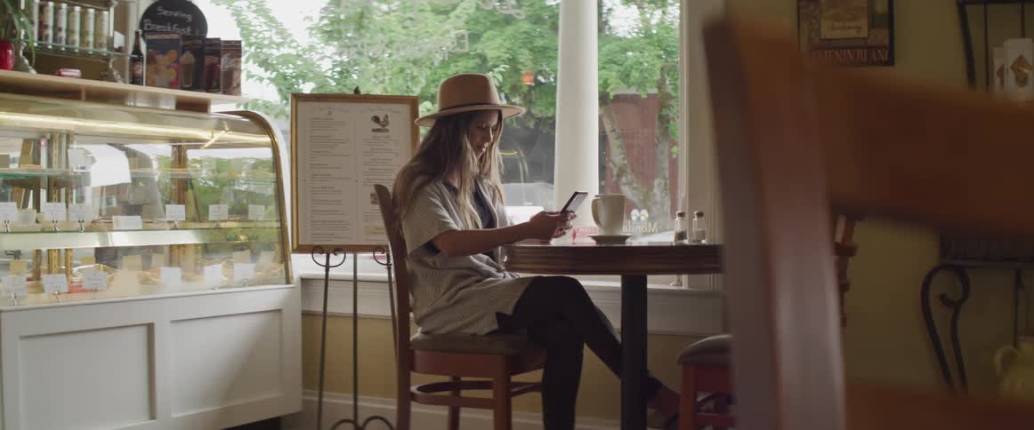 A woman wearing a straw hat is sitting at a round table near the window in a bakery while scrolling on her tablet