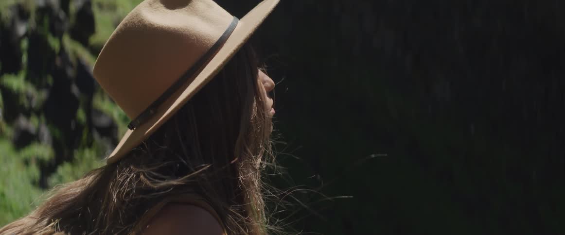 Young woman with long hair and a cowboy hat stares off into the distance in a forest on a sunny day