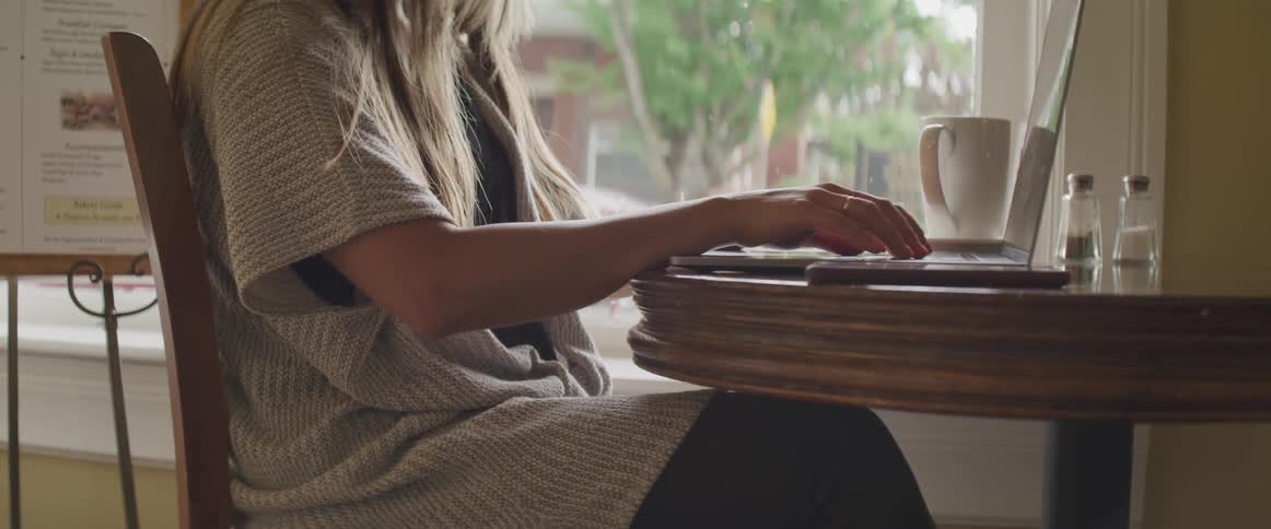 A woman is sitting at a wood table typing on her laptop with a coffee cup nearby
