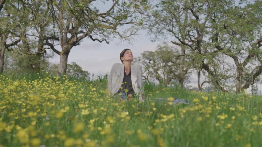 A woman sits in a field of yellow flowers with her eyes closed and face tilted up towards the sun