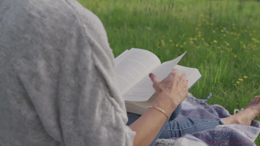 Woman sits on blanket in meadow reading a book