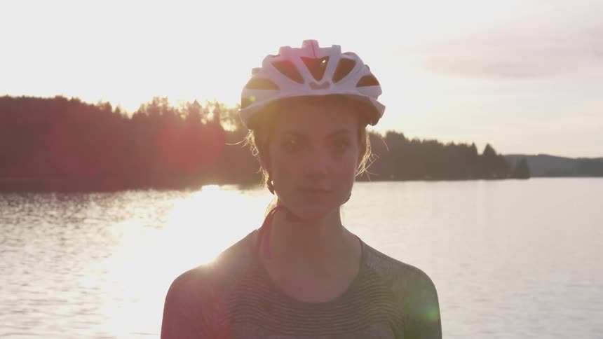 A woman wearing a white bike helmet stands in front of a tranquil lake at sunset