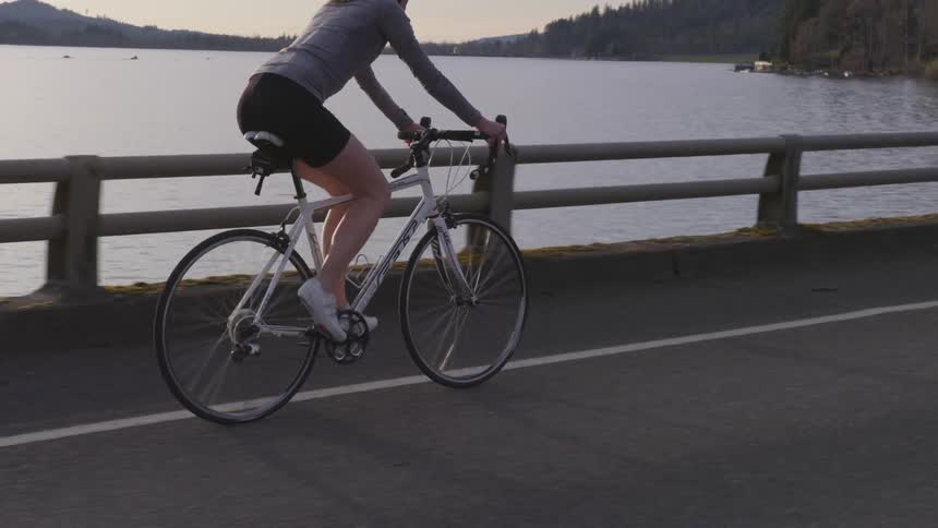 A woman rides a bicycle on a road along the water at sunset
