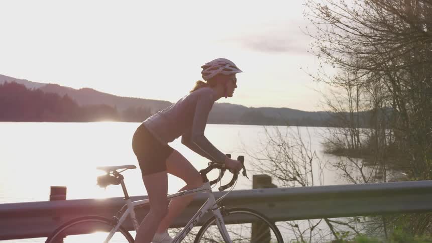 A woman in a white helmet bicycles next to a lake at sunset
