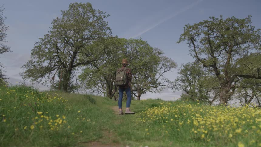 A woman pauses in front of some trees while on a dirt trail as she is backpacking and hiking up a grassy hill
