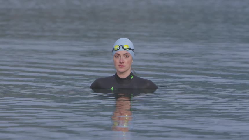 A woman sits in the water wearing a full body swimsuit swim cap and goggles