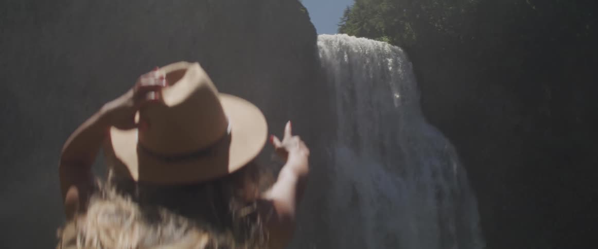 A white woman wearing a brown hat points at a waterfall