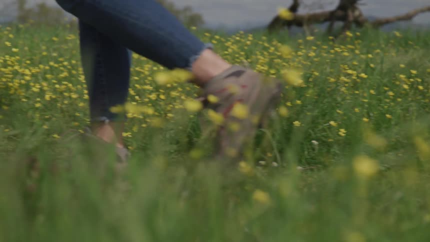Woman wearing sneakers and denim walks on the path through the field with yellow flowers during daylight