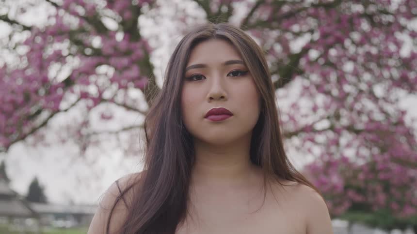 A woman with bare shoulders stands outside with a pink blossoming tree behind her