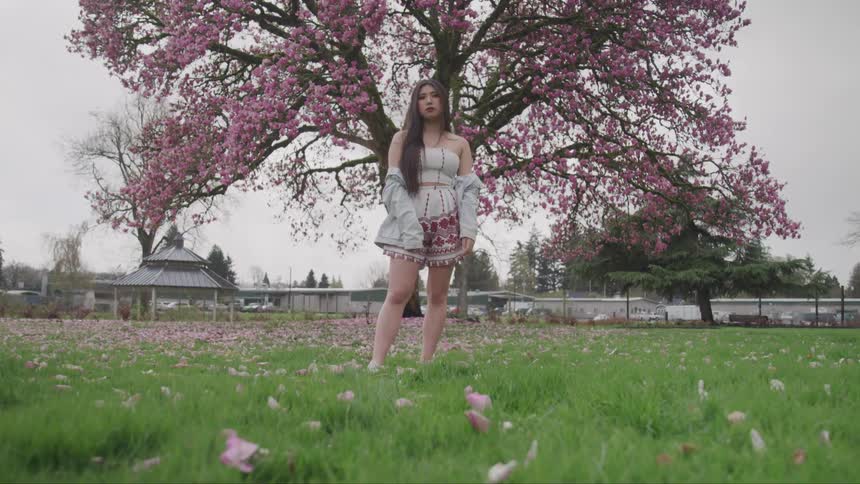 Young woman stares into the camera as she stands in front a cherry tree in field of grass covered with fallen pink petals