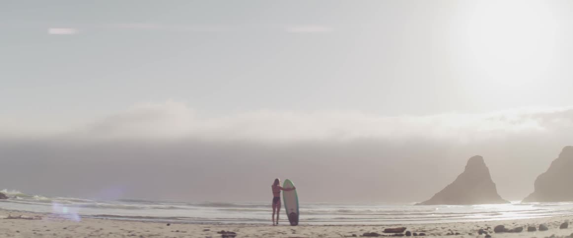 A woman holds her surfboard and stands on the beach while looking out to sea