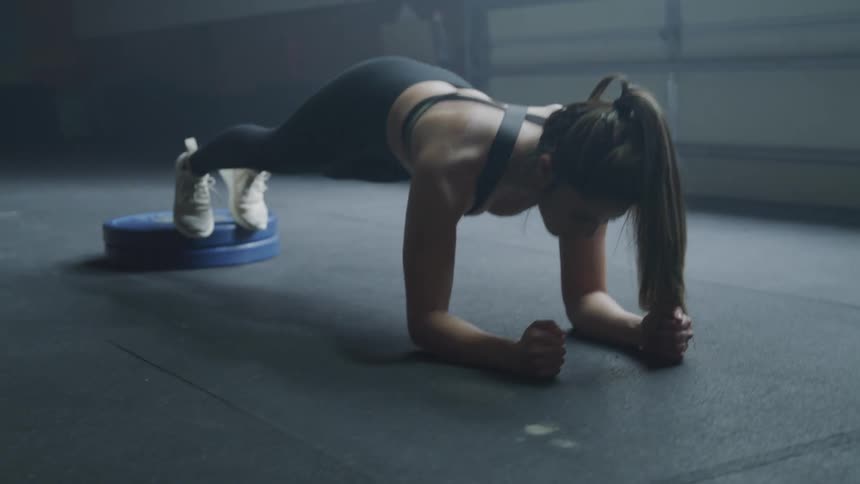 Muscular young woman does planks exercise in the dim light in gym