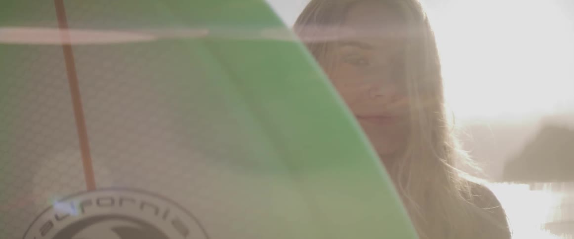 A female holds a surfboard and looks into the camera at sunset