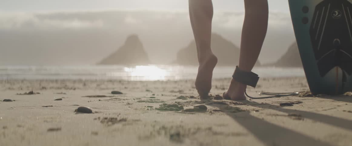 A woman standing next to a surfboard on the beach is drawing in the sand with her toe in front of the ocean
