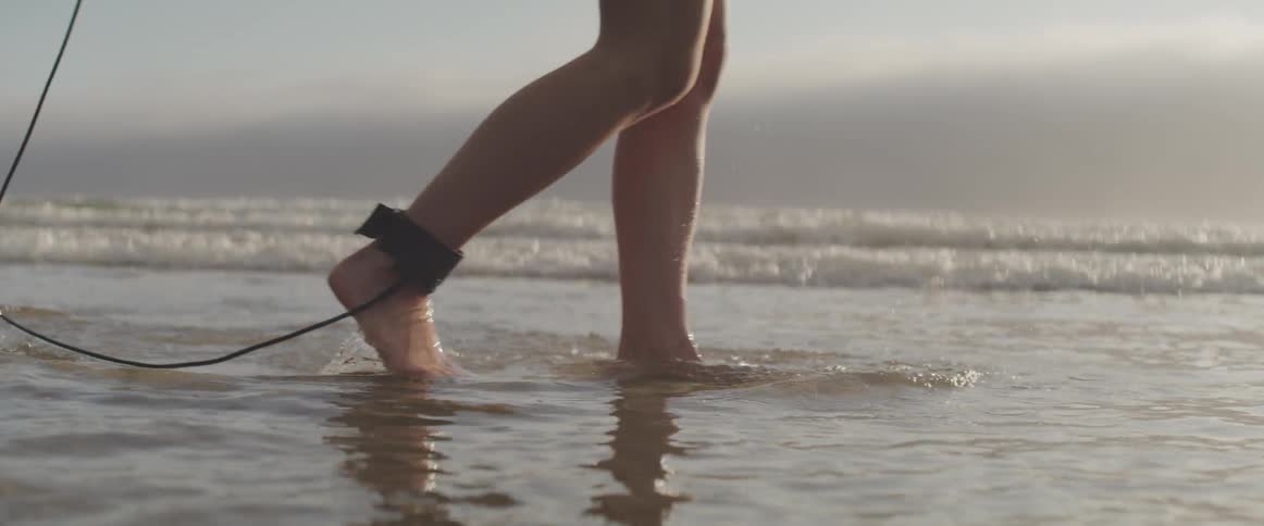 A person walks through the ocean water with a surfboard strapped to their ankle