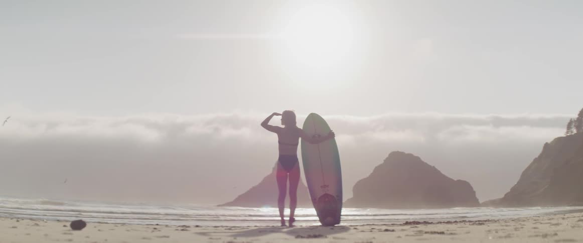 A woman in a bathing suit and holding a surfboard on the beach looks out over the water