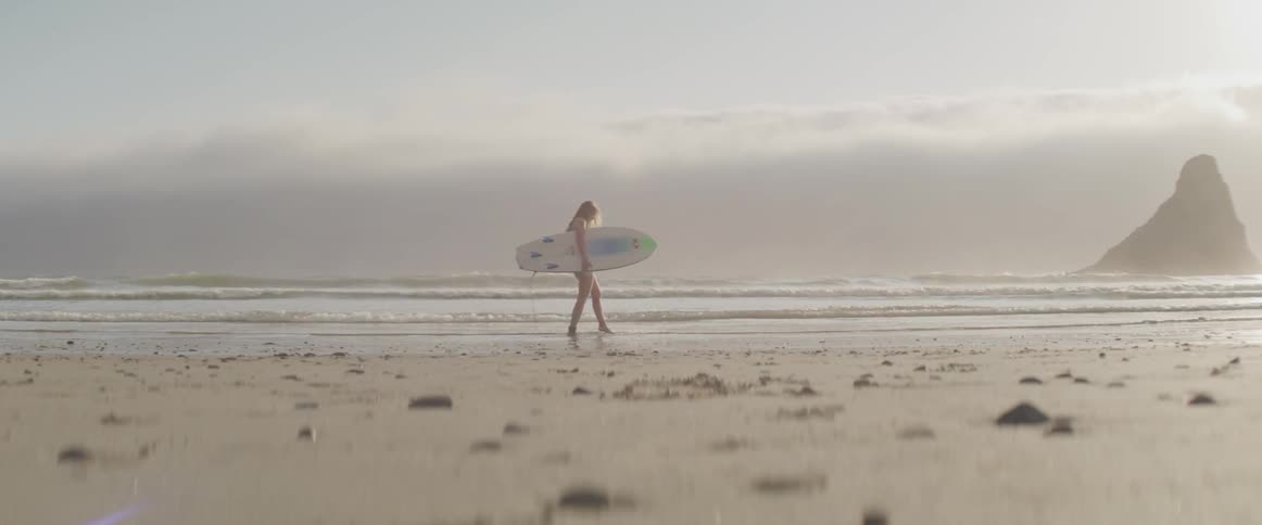 A young white girl holding a surfboard walks along a sandy beach and kicks the water