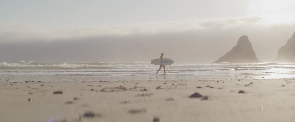 A woman carries a surf board along a beach