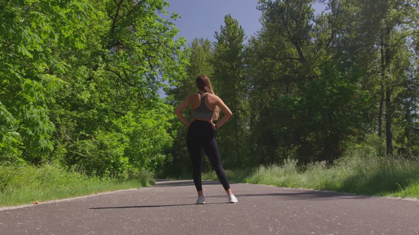 Athletic woman wearing running pants and sports bra stands in the middle of a paved road surrounded on either side by forest trees on a sunny day