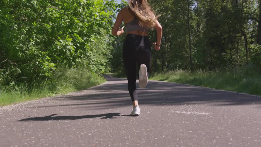 A white female with long blonde hair runs on a paved path through a park on a sunny day