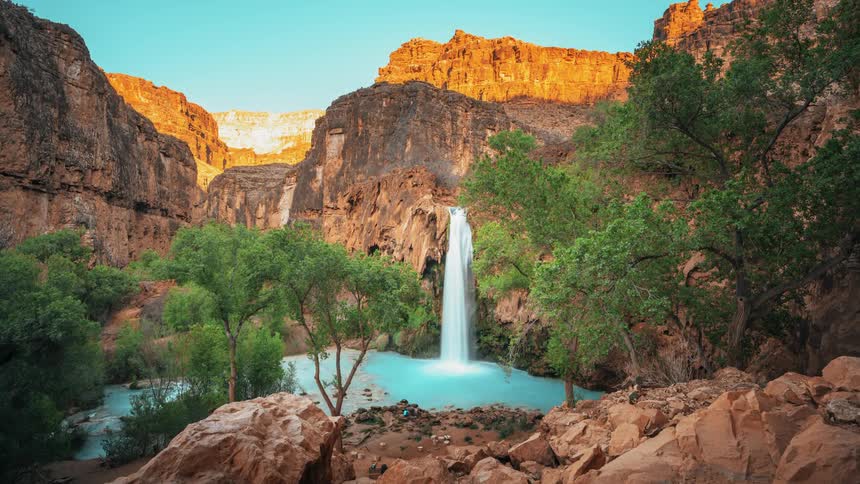 A waterfall flows over a red rock cliff into a pool of turquoise water below