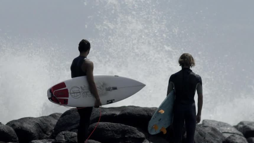 Men holding surfboards while waves crash against the boulders they are standing on