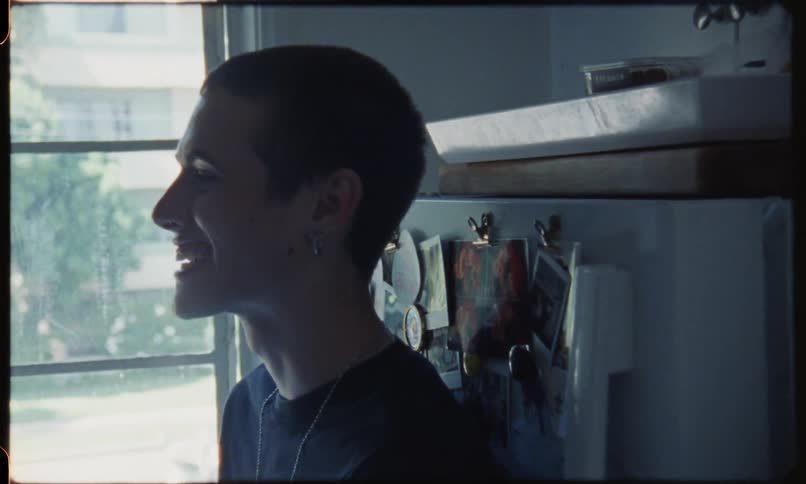 A white teenager is talking and laughing with someone while leaning up against a fridge in a kitchen